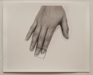 A photo of a black and white photograph with a hand, palm facing down, on paper. A square is drawn on the tip of the index and middle fingers and on the paper.