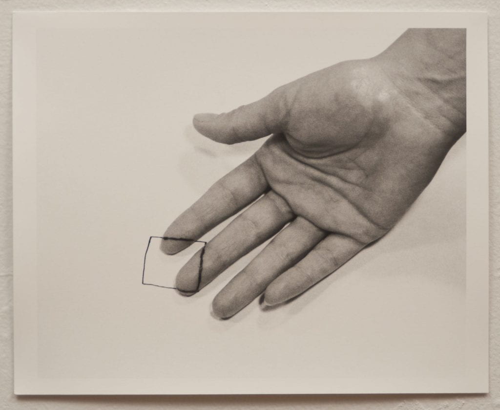 A photo of a black and white photograph with a hand, palm facing up, on paper. A square is drawn on the tip of the index and middle fingers and on the paper.