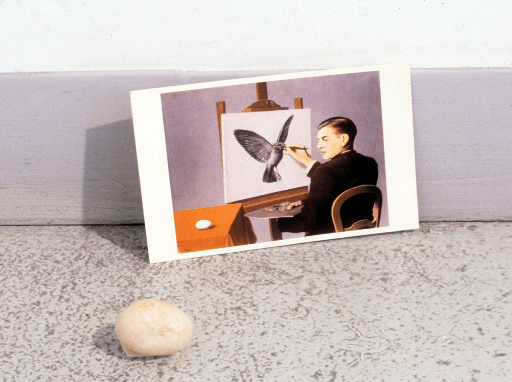 A postcard of a man in a black suit painting a gray bird in flight. He stares at an egg to his left on an orange table. A stone shaped like an egg is placed on the floor to the left of the postcard.
