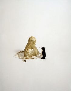 A photo of a gold lamp in the shape of Christ and a wooden penguin gazing closely at each other against a white background.