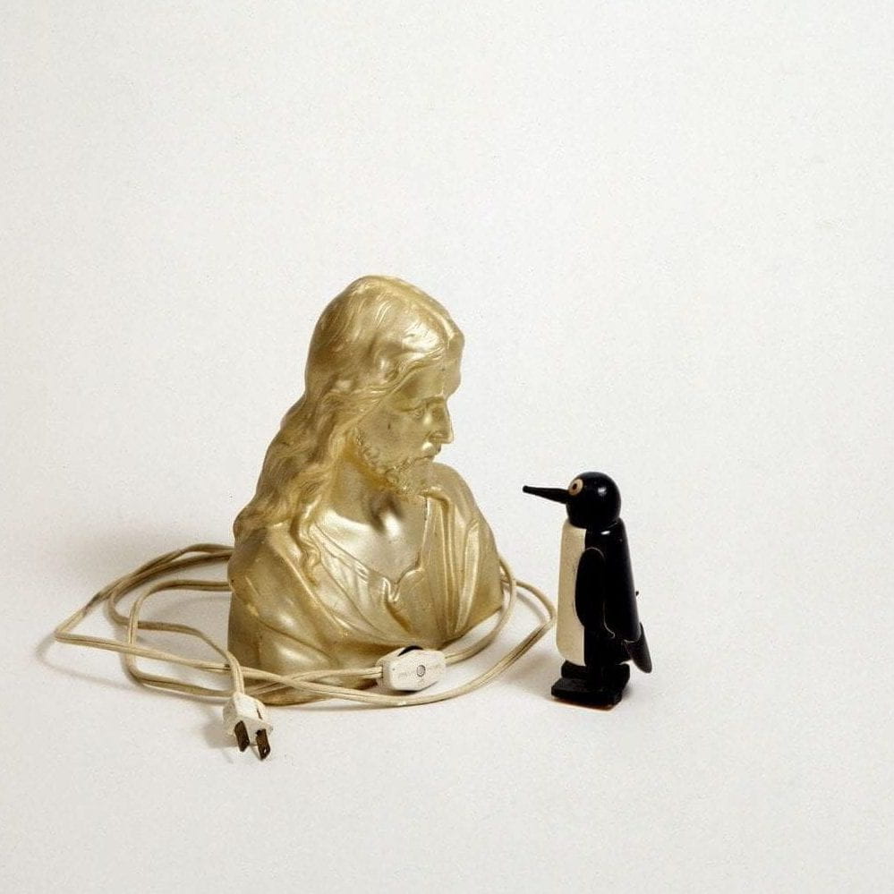 A photo of a gold lamp in the shape of Christ and a wooden penguin gazing closely at each other against a
white background.