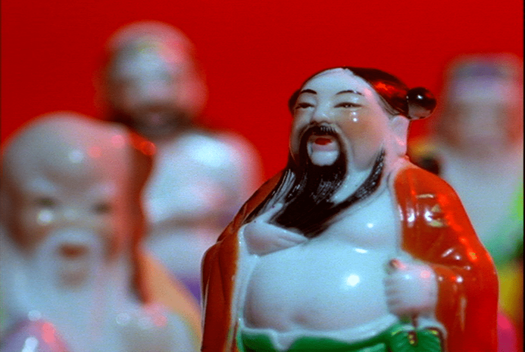 A video still of a pale, portly figurine with long black hair and beard in an open red robe. Similar figures fill the background.