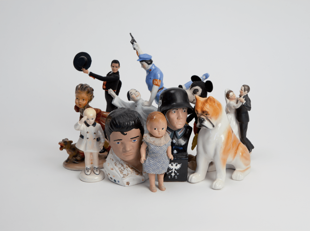 A photograph of a group of eleven figurines including children, a Nazi soldier, a dancing couple, a porcelain dog, and Mickey Mouse.