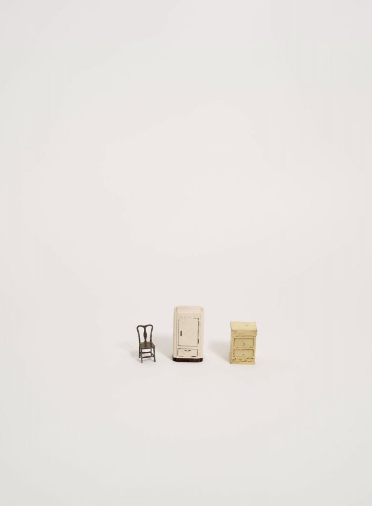 A photograph of miniature furniture (a brown chair, white refrigerator, and beige end table) lined up on a white background.
