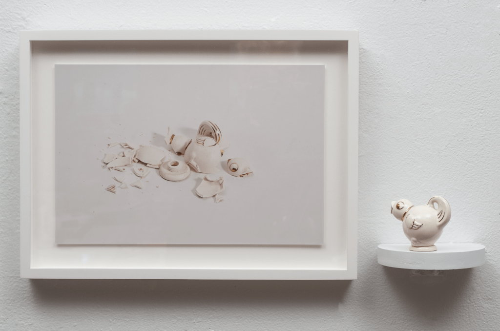 A framed photograph of a broken porcelain white bird. Displayed to the right of the photograph is the bird itself on a shelf, unbroken. 