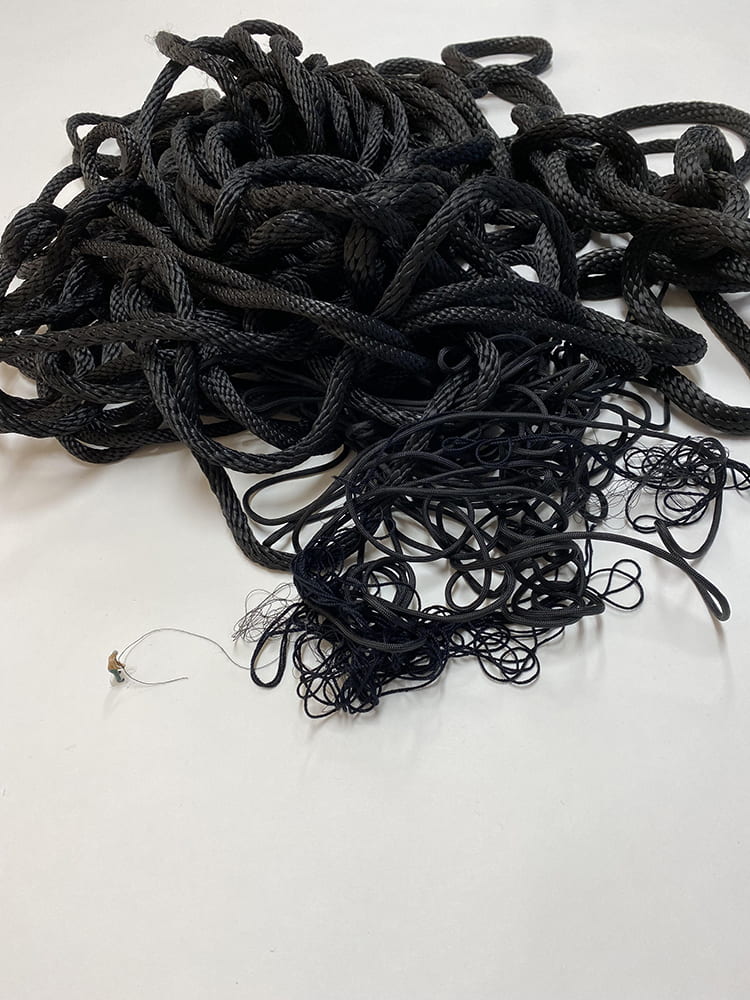 A photograph of an installation of a miniature toy man on a white pedestal with a black string in his hands. The string connects to a large tangled pile of black rope. 