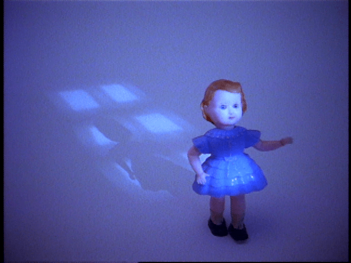 A video still of a blonde doll in a blue dress on a dark bluish background. A shadow of the doll and a window appears behind the figurine. 