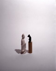 A photo of a porcelain woman in a robe looking into the eyes of a short black bear figurine against a white background. The bear is atop a brown cylinder to reach the woman at eye level.