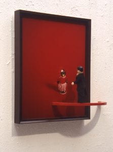A framed print of a penguin in a red dress against a red background. A suited toy man looks at the penguin from a semicircle platform attached to a wall.