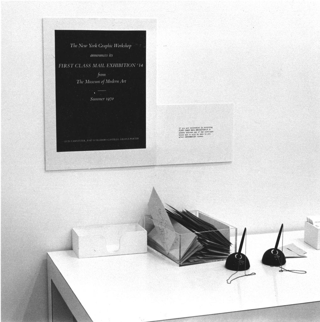 A photograph of a white desk with a clear plastic box of envelopes and two black pens, signage mounted on the wall. 