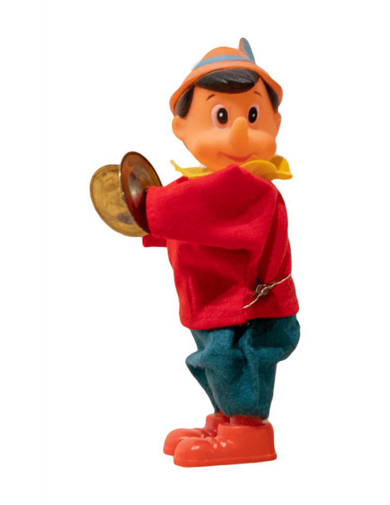 A photo of a Pinocchio-like figure holding cymbals and staring at the camera. He has a pointy nose and wears a red shirt, blue pants, and feathered cap.